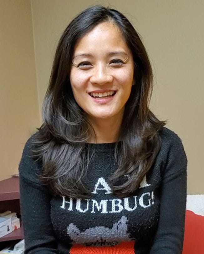 Woman with long dark hair smiling, wearing a black sweater that says "BAH HUMBUG!" and sitting indoors.