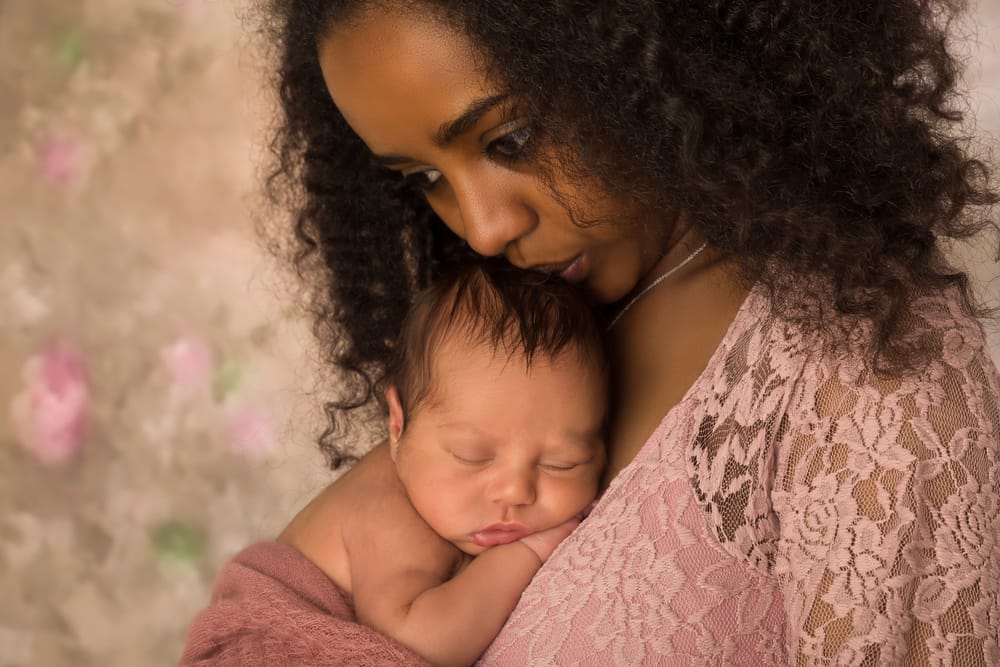 A woman in a pink lace robe lovingly holds and kisses a sleeping newborn baby on her chest.