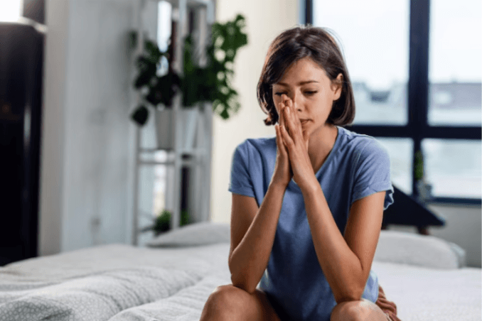 A woman sits on a bed with her hands clasped, appearing stressed or deep in thought, as she reflects on healing emotional trauma.