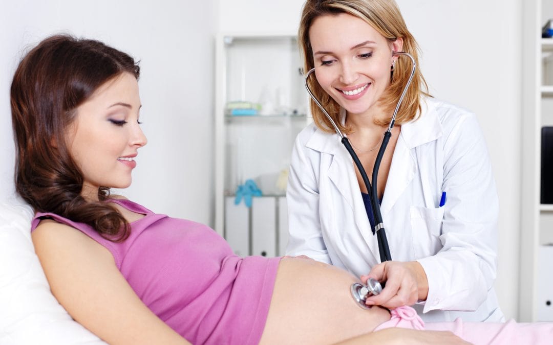 A doctor and a Fertility Acupuncturist work together as the doctor uses a stethoscope to check a pregnant woman’s belly during a prenatal exam.