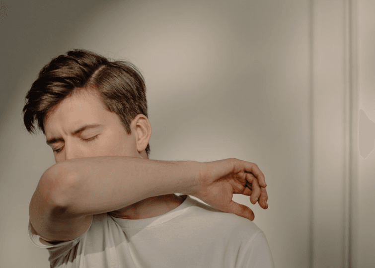 A person in a white shirt demonstrates good hygiene by sneezing or coughing into their elbow against a neutral background, which supports wellness practices often encouraged alongside acupuncture.