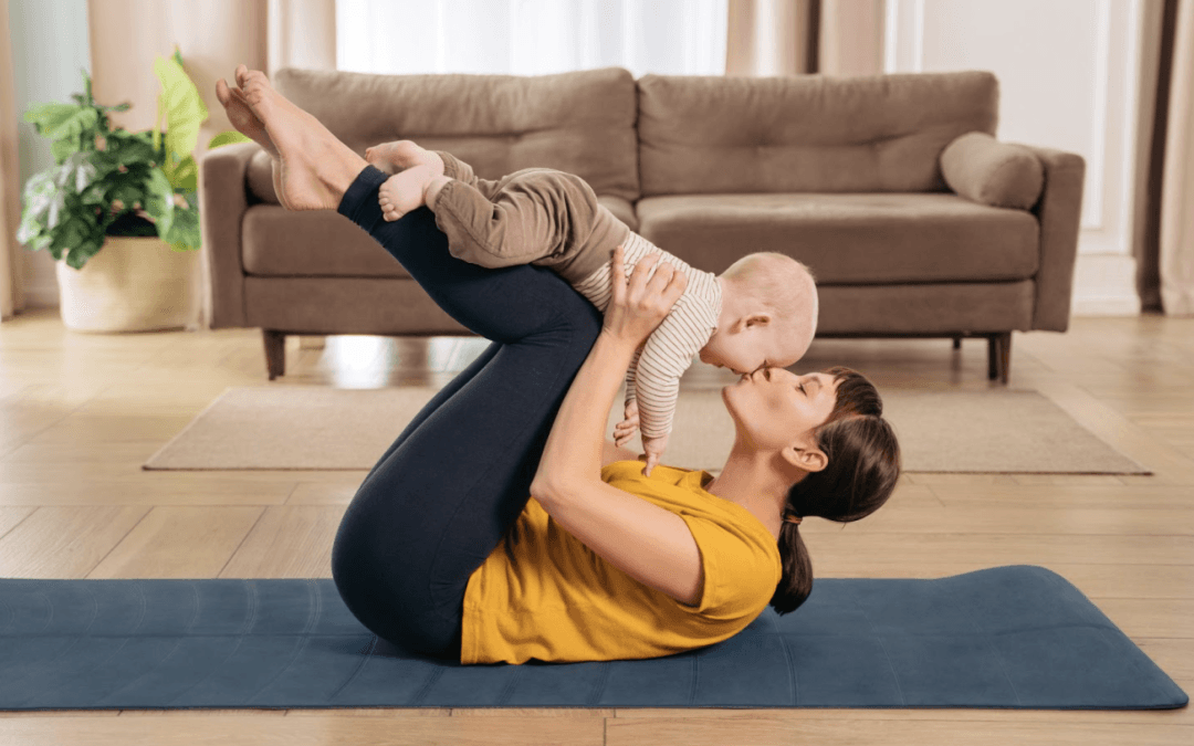 A woman lying on a yoga mat playfully lifts a baby above her, both smiling in a cozy living room. Postpartum Recovery