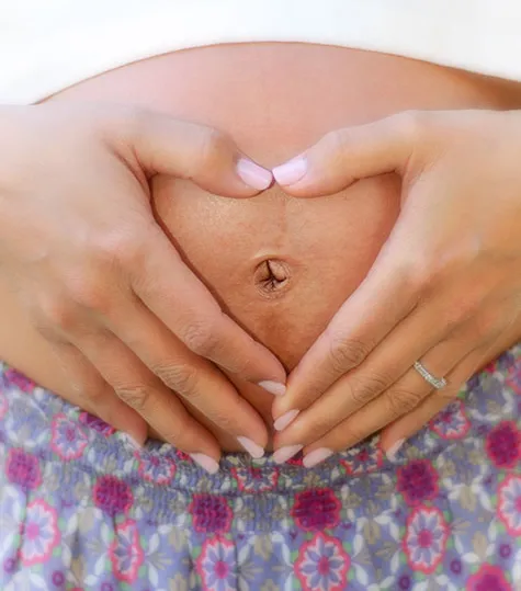Pregnant person wearing a ring forms a heart shape on belly with hands, above floral-patterned skirt.